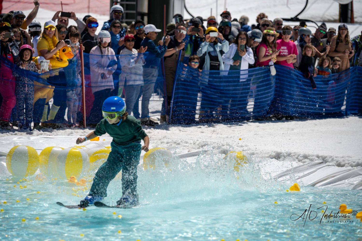 Skier in green outfit performs water crossing stunt at winter resort event with spectators.