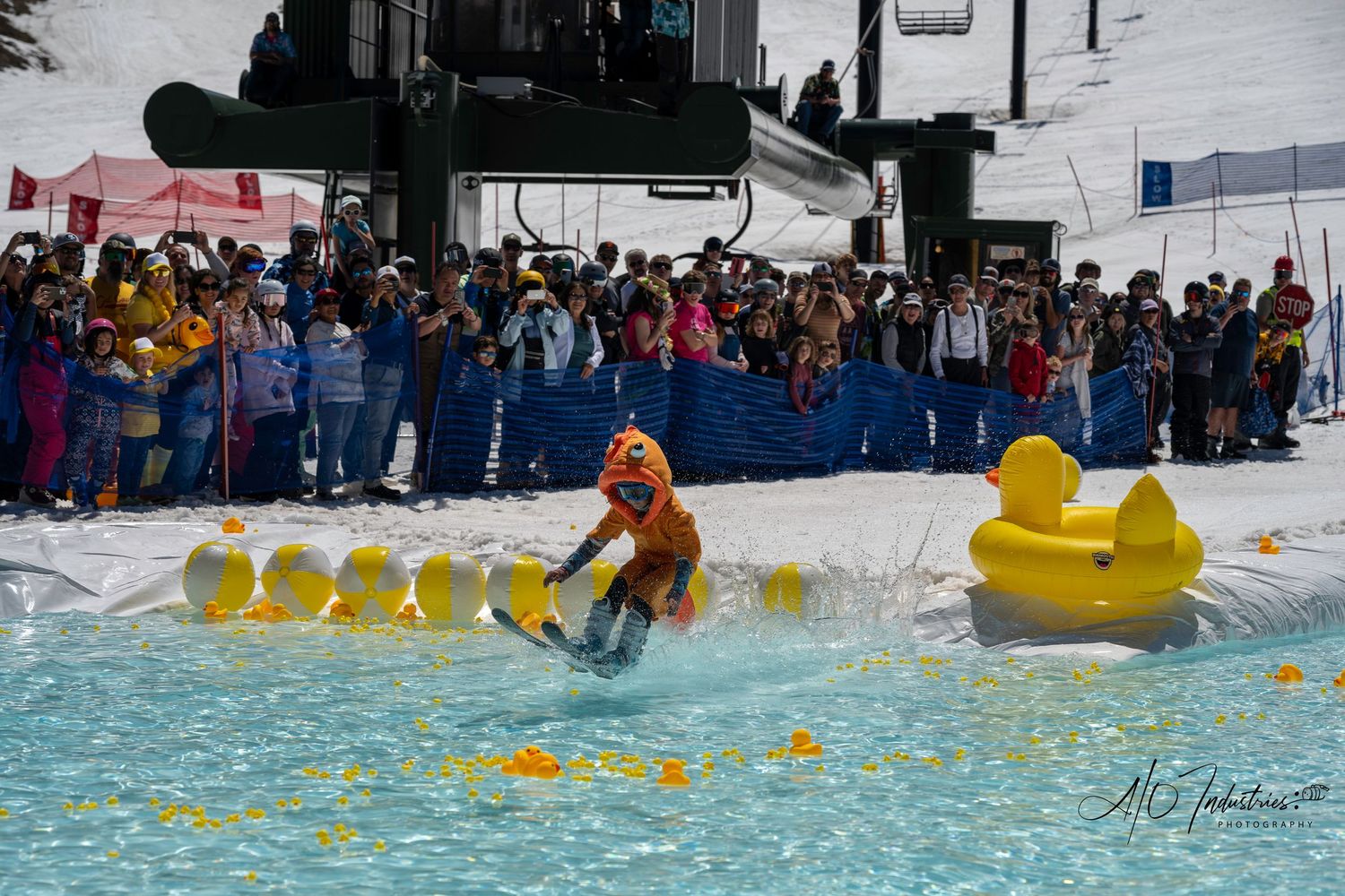 Skier in orange outfit crosses water pool with giant rubber duck during spring skiing event.