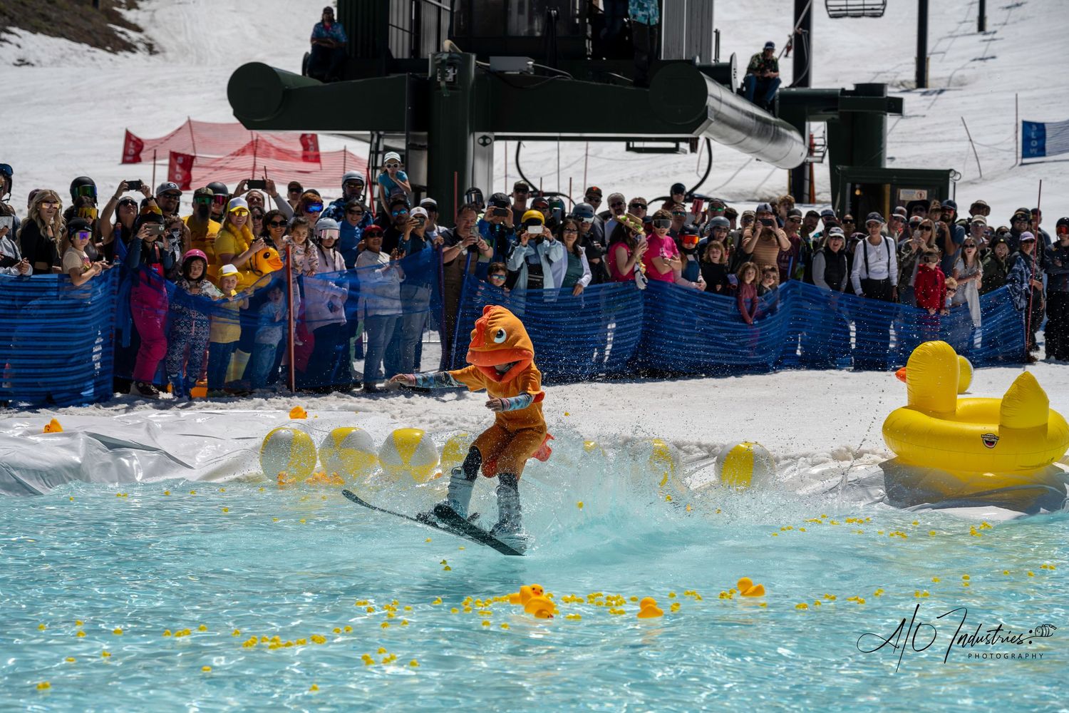 Skier in bright orange navigates water crossing with yellow duck decorations and crowd watching.