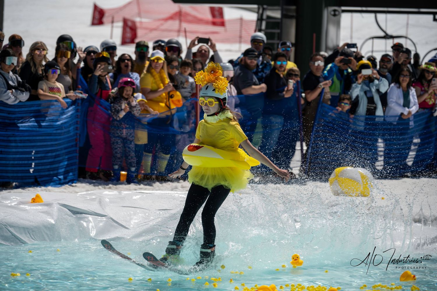 Skier in yellow outfit jumps across water while crowd watches from behind barriers.