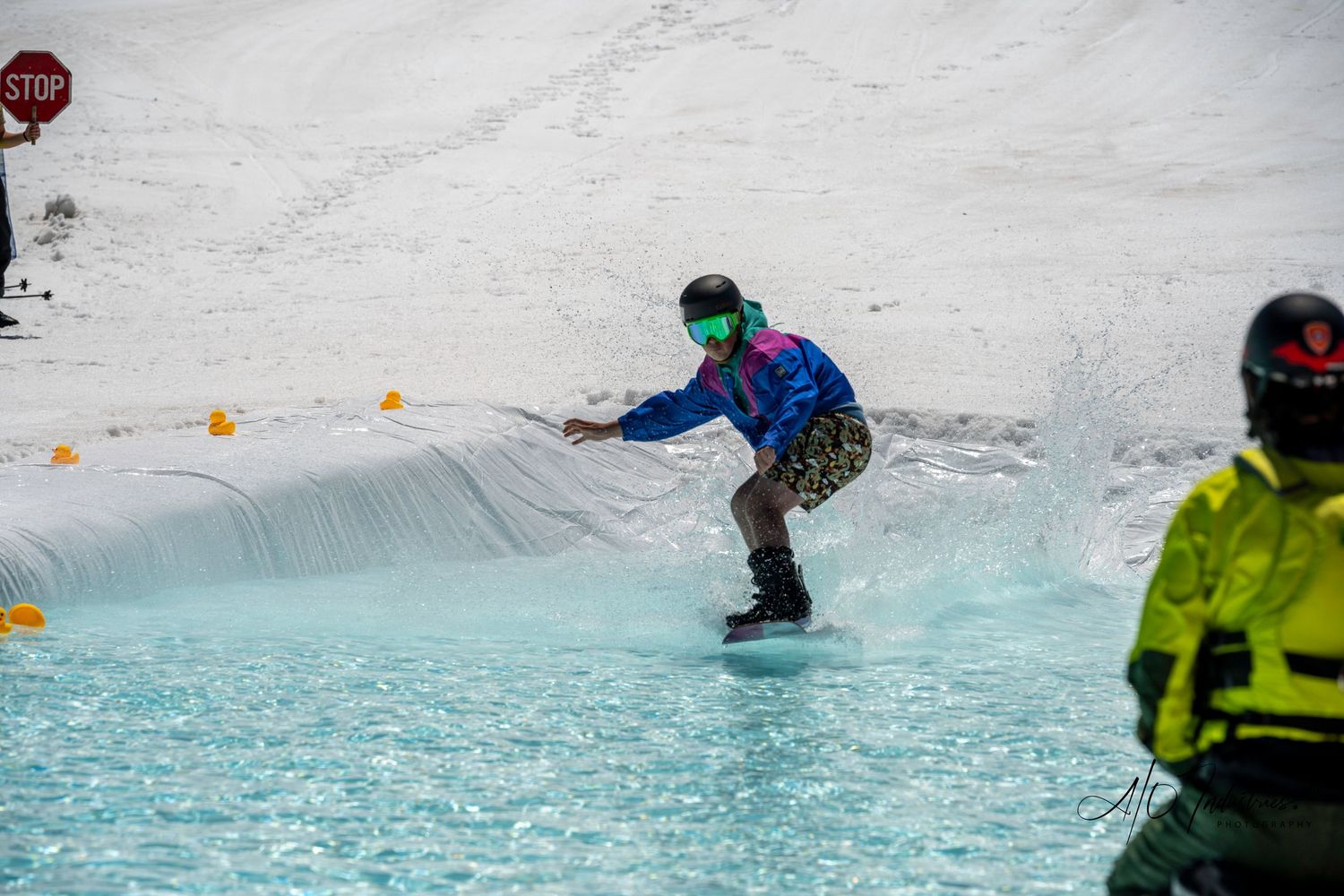 Snowboarder crosses blue pond with yellow buoys during spring skiing event.