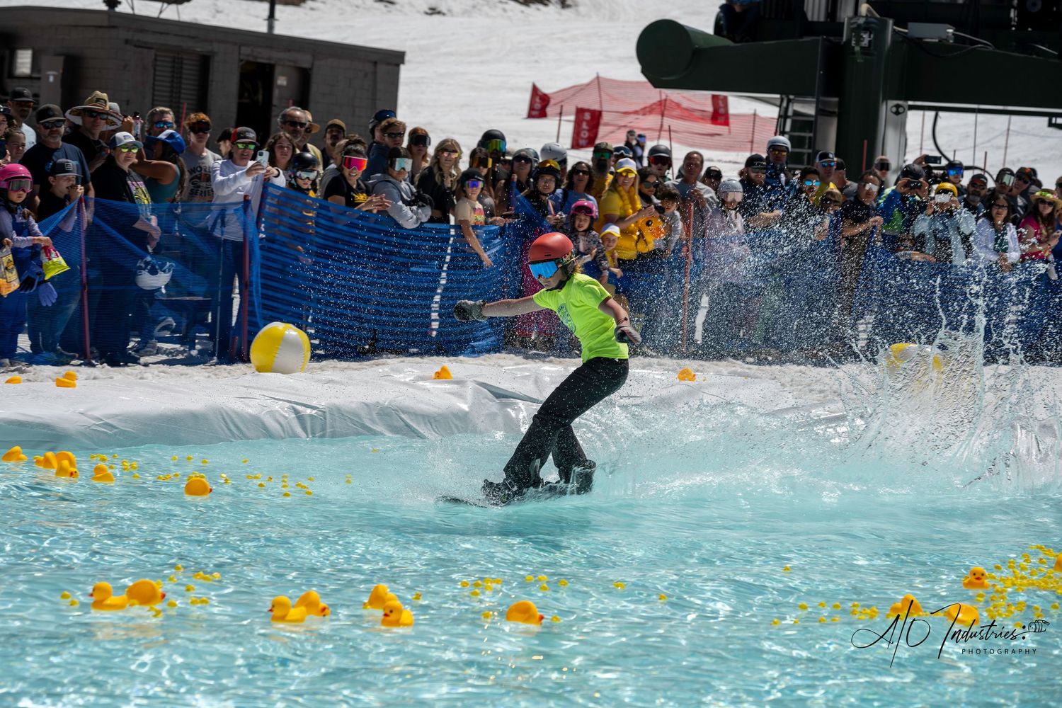 Snowboarder skims across bright blue pool while crowd watches from behind safety barriers.