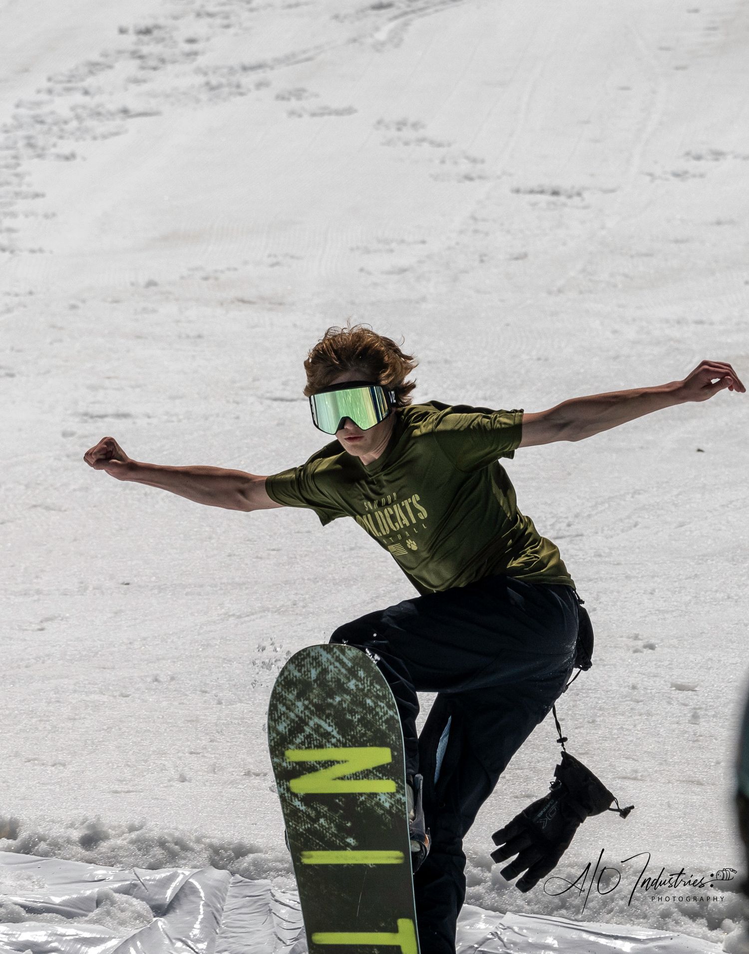 Snowboarder in green shirt strikes dramatic pose on snowy slope with arms spread wide.