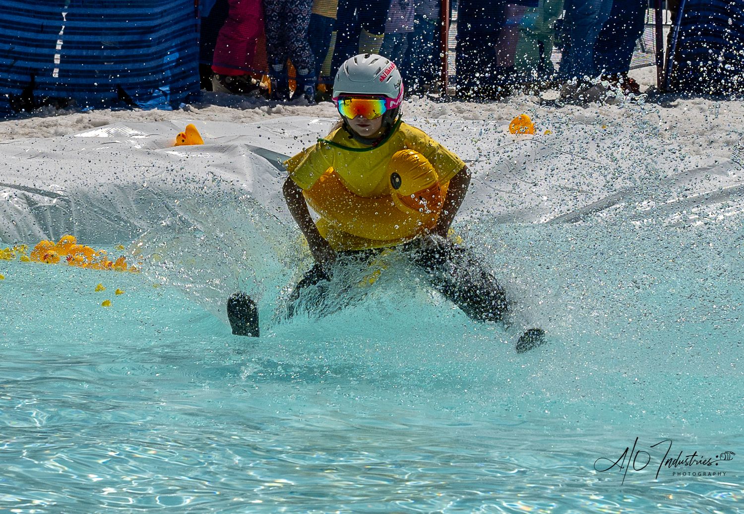 Snowboarder in yellow jacket splashes through water during pond skimming competition.