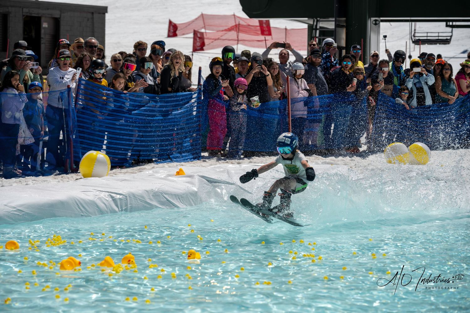 Crowd gathers around water pool to watch snowboarders attempt pond skimming tricks.