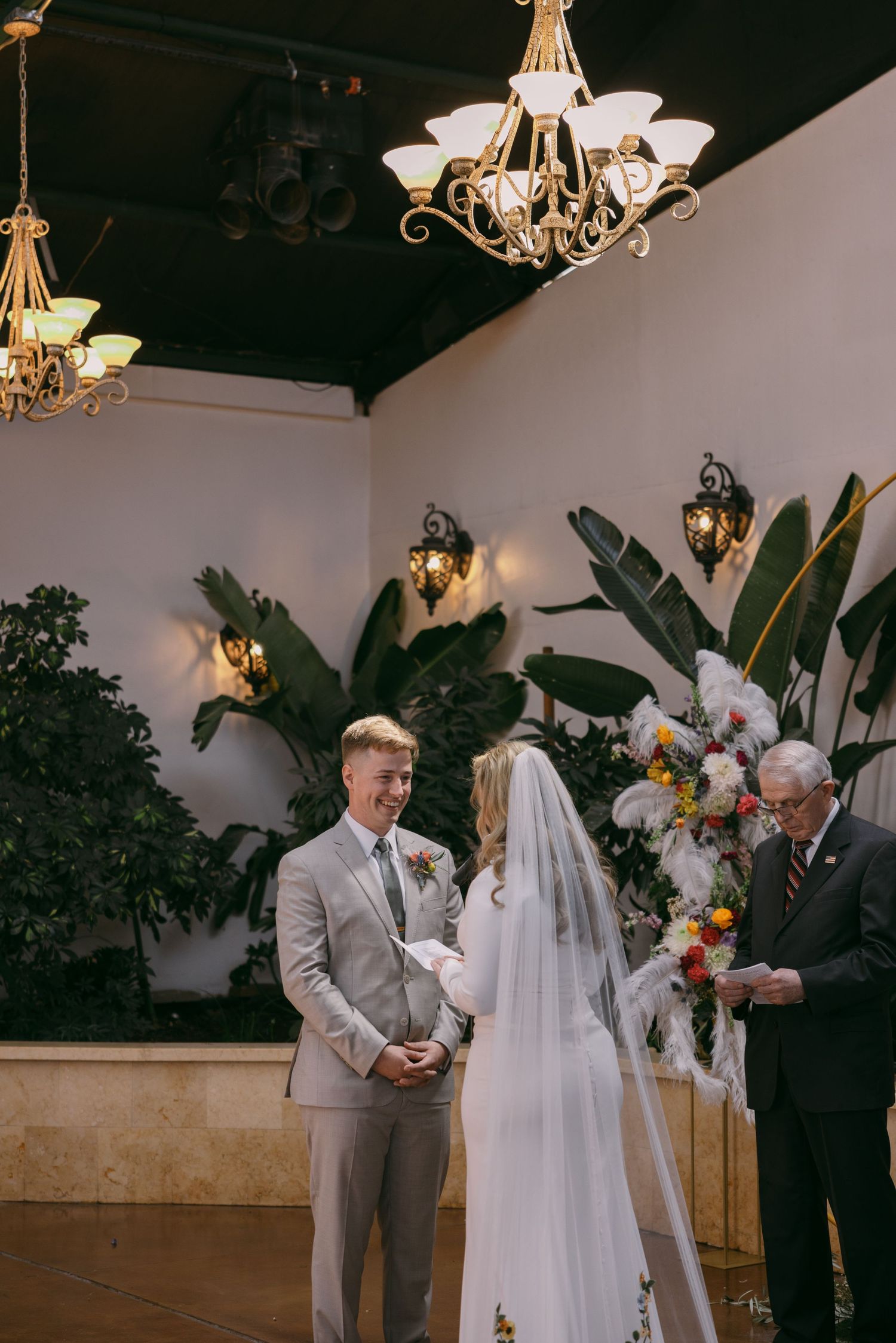 An indoor wedding ceremony with tropical plants and elegant chandeliers in the background.