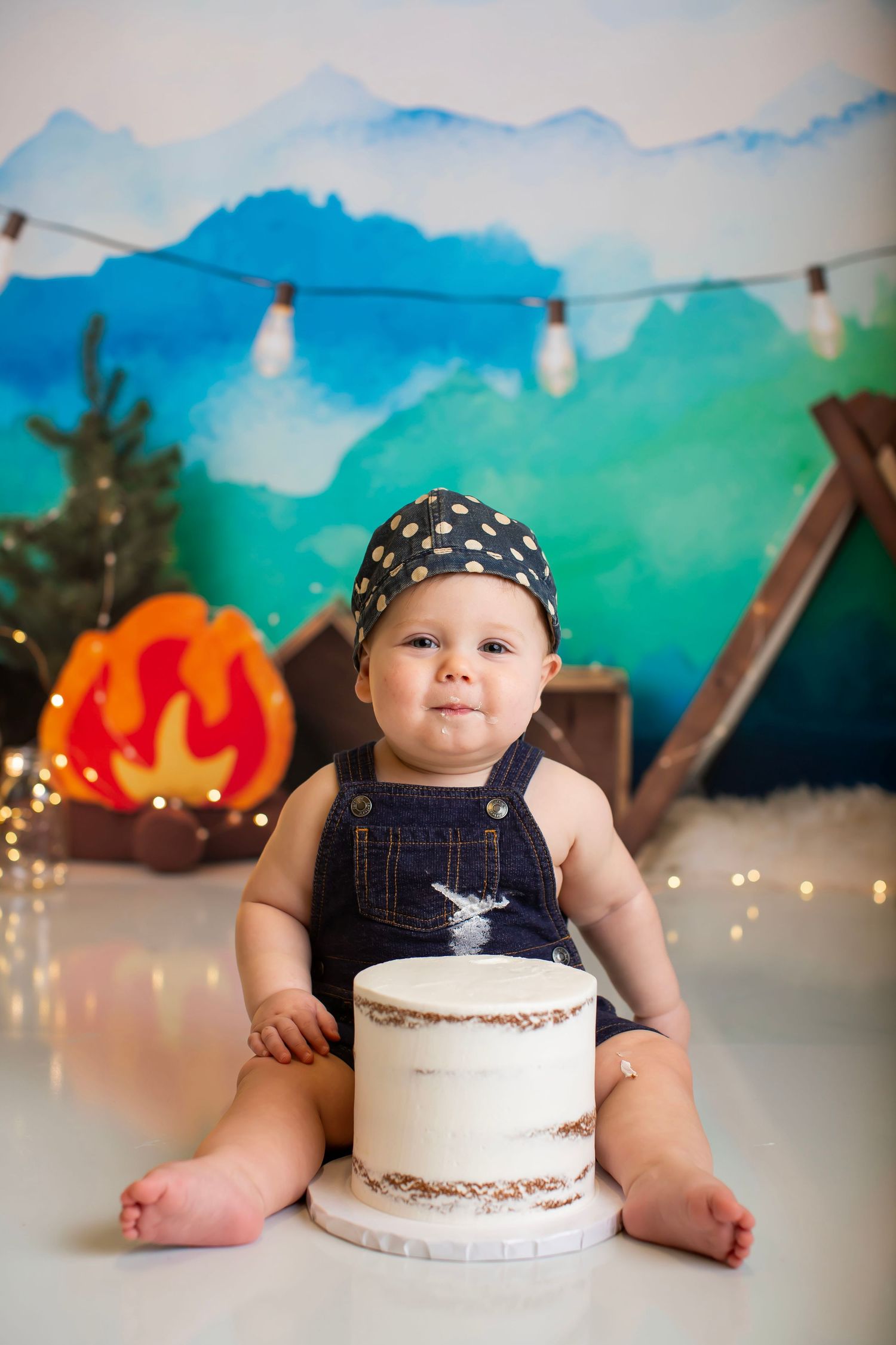 Baby in overalls sits with cake against mountain backdrop with string lights.