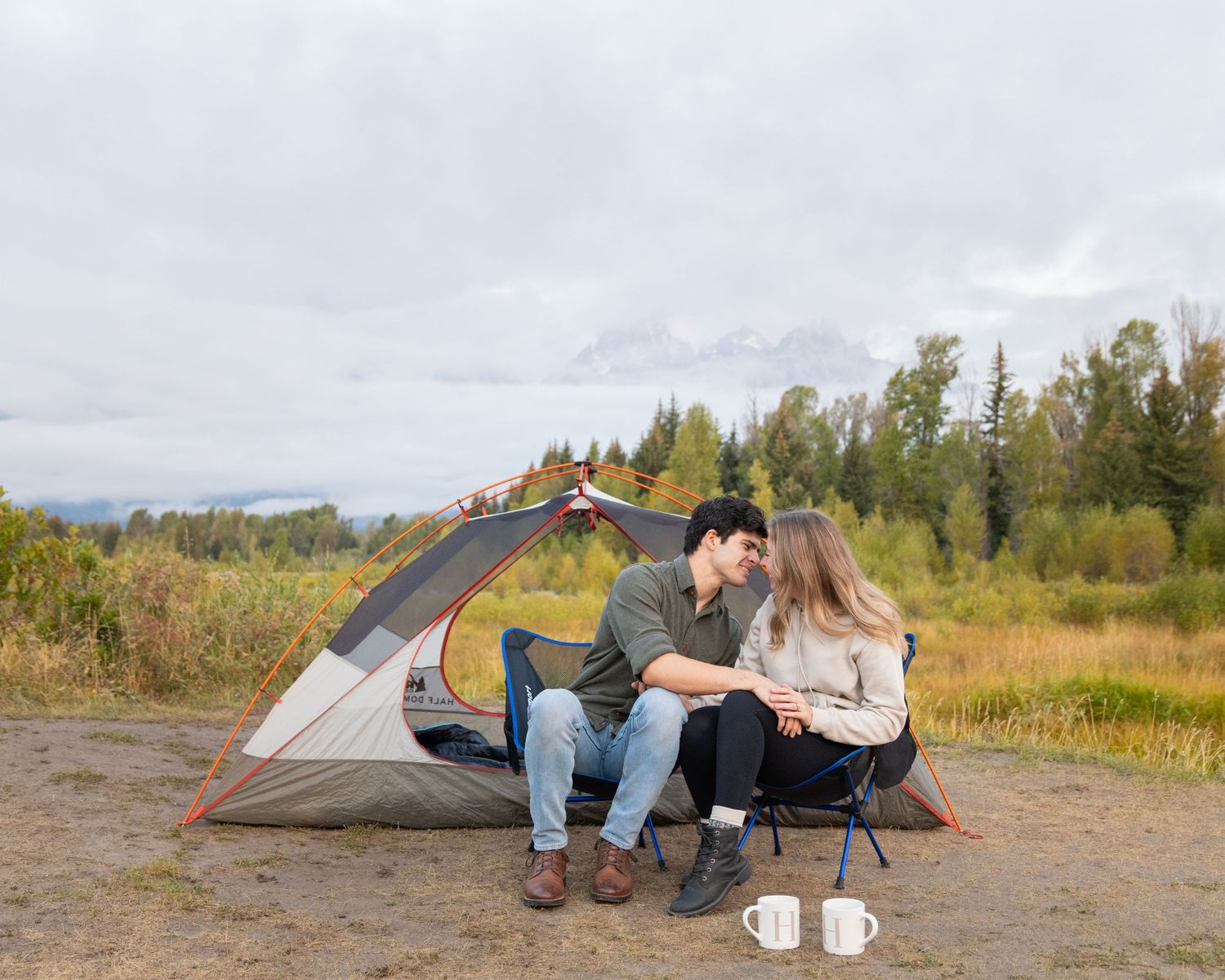 Couple sitting outside camping tent in wilderness with coffee cups on cloudy day.