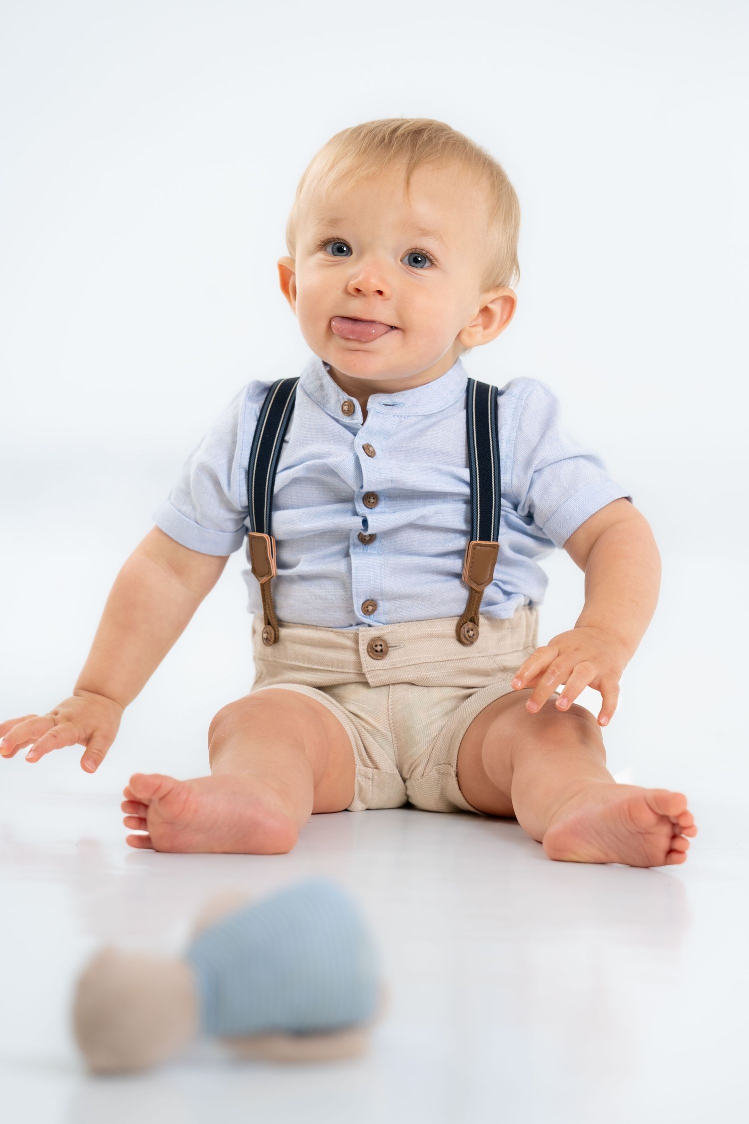 Baby in light blue shirt and suspender shorts sitting on white floor.