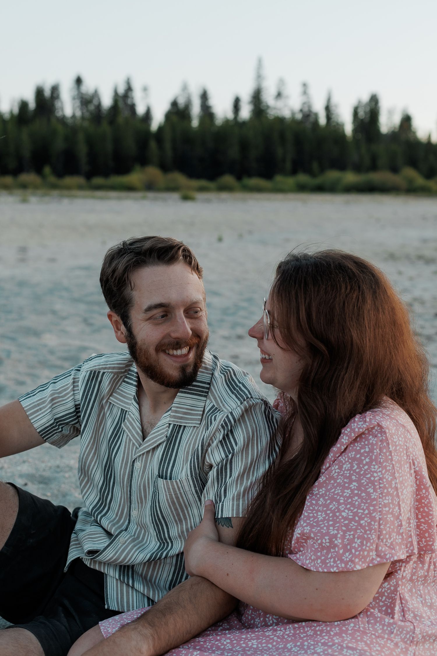A couple shares a romantic moment by the lake in Central Oregon with pine trees in the background during sunset.