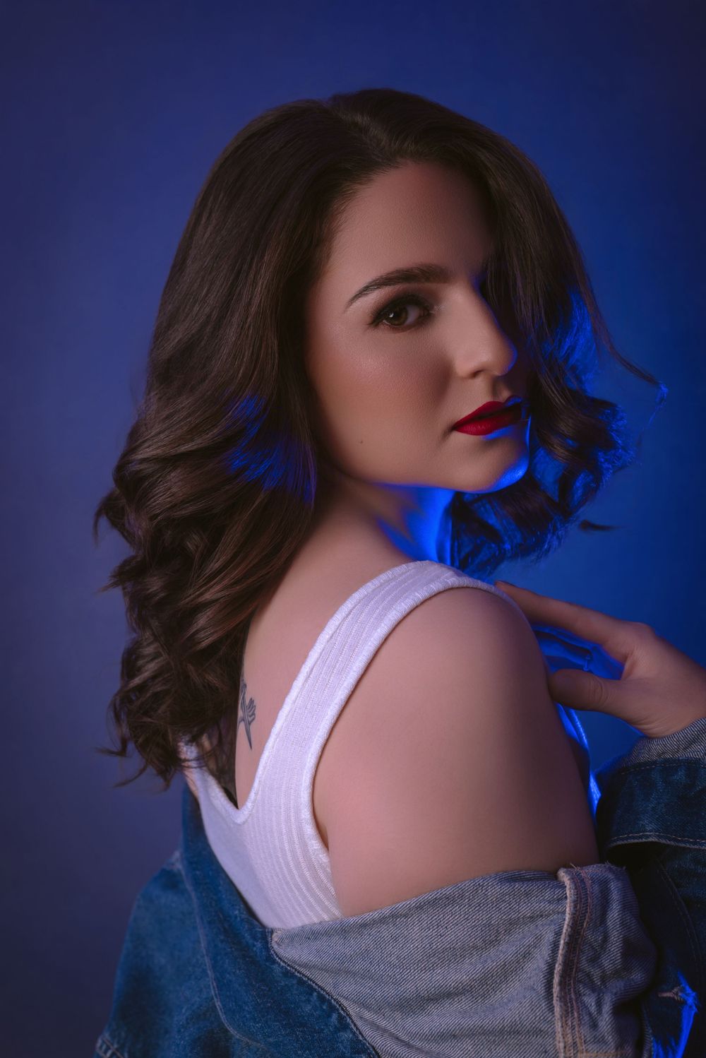 Artistic beauty portrait with white top and denim jacket against blue backdrop.