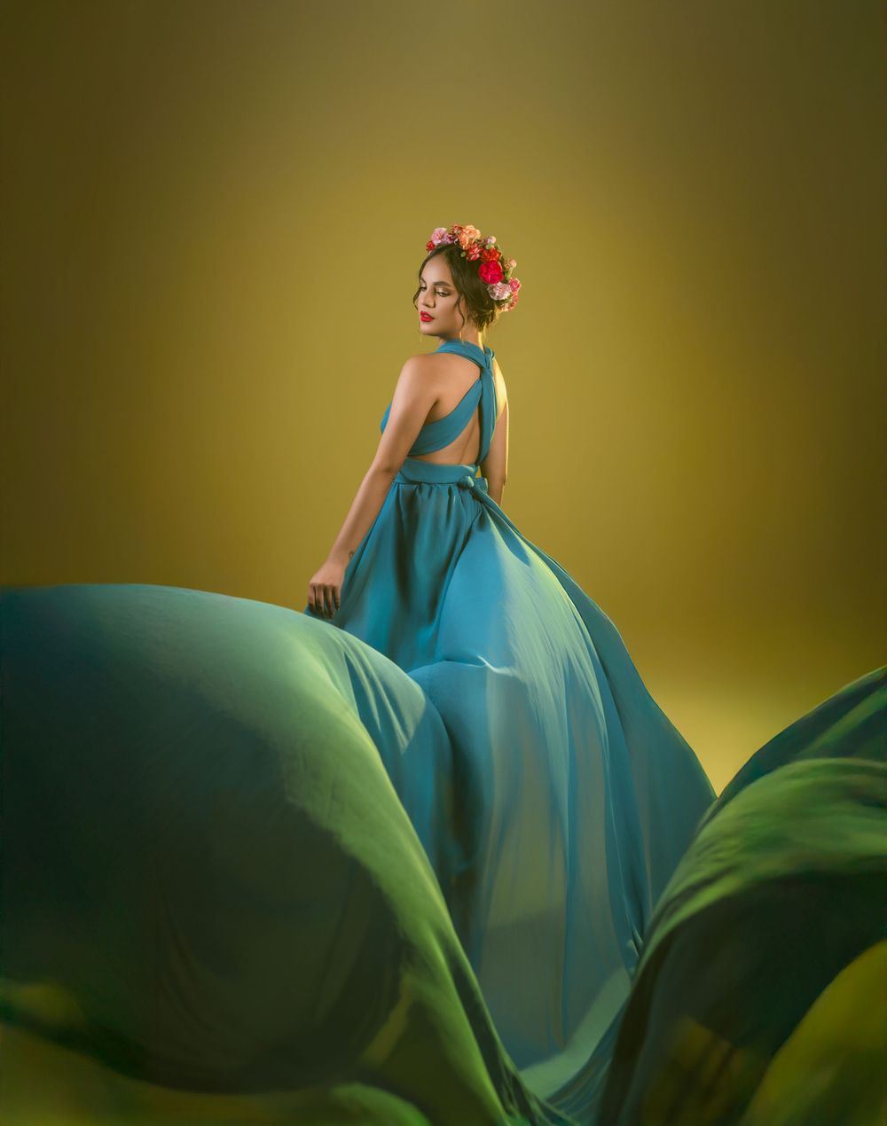 A flowing turquoise gown and floral headpiece create a dramatic fashion portrait against a yellow backdrop.