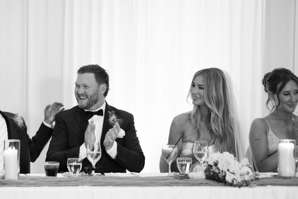 Black and white wedding reception photo of laughing bride and groom seated at head table with candles and flowers.