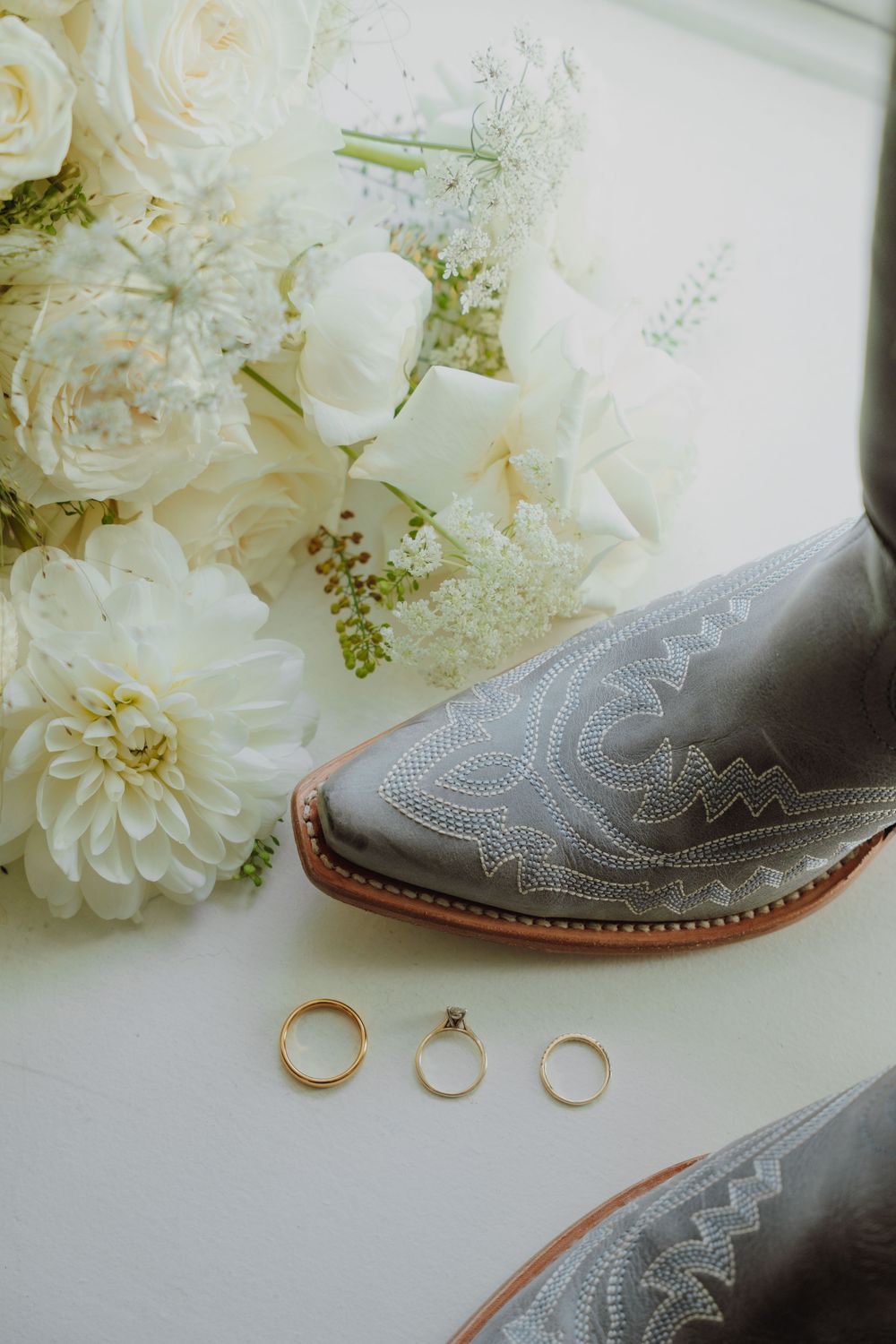 Gray cowboy boots beside gold wedding rings and a lush white floral bouquet with roses and dahlias on a white surface.