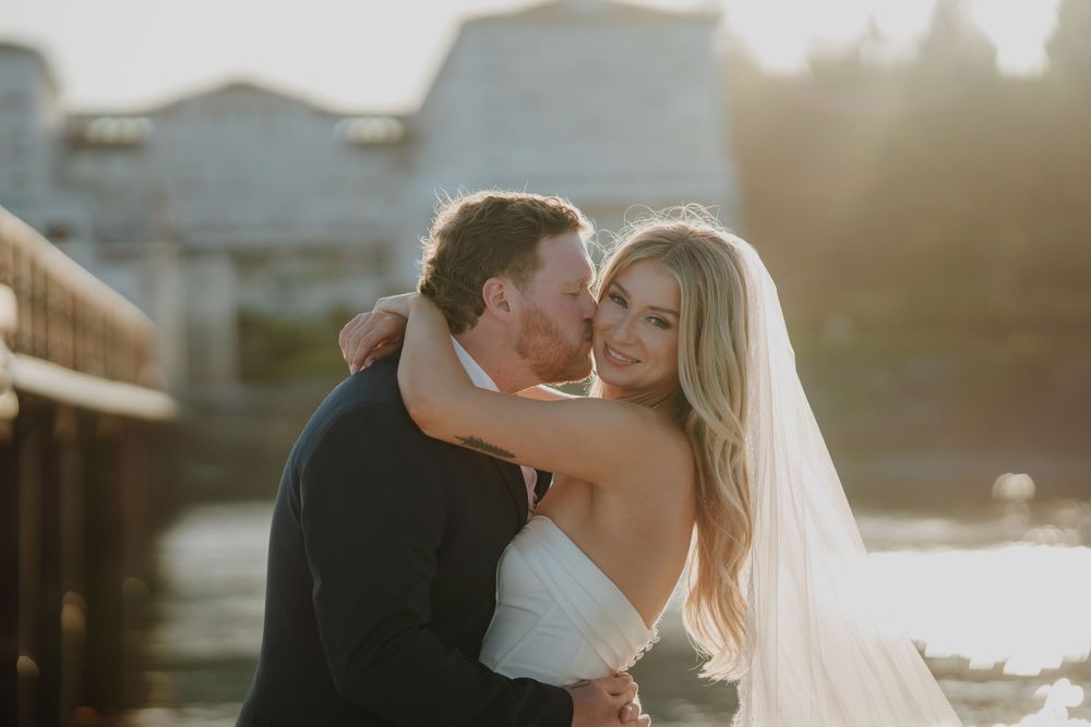 Groom kissing bride on cheek at waterfront wedding, golden hour sunlight, bride in white strapless gown with veil.