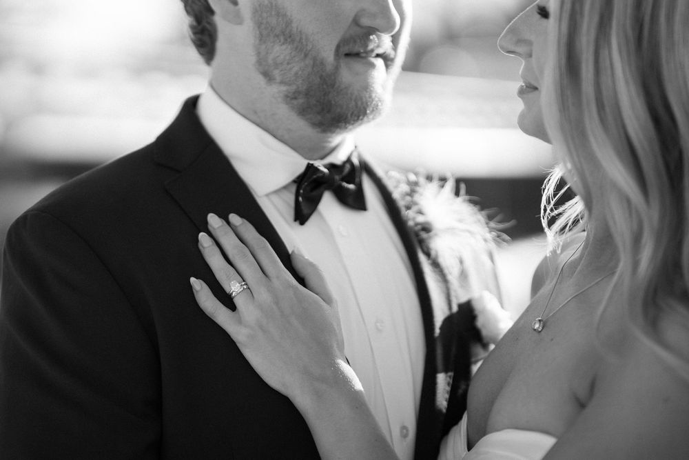 Black and white wedding photo of bride placing hand on groom's chest, showcasing engagement ring, bow tie, and formal attire.