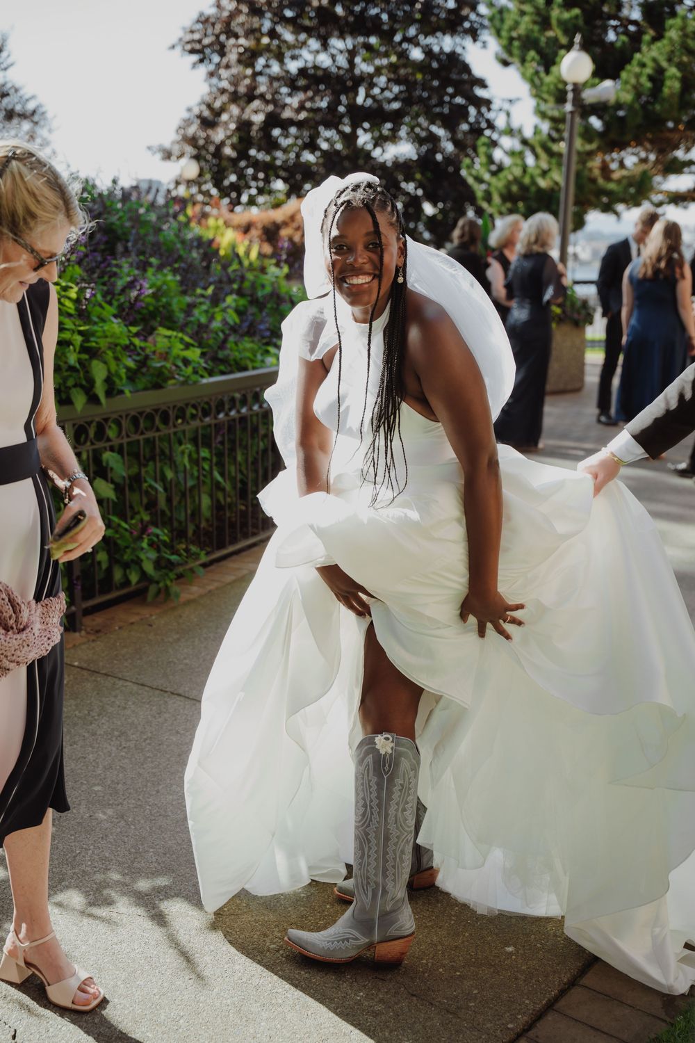 Smiling bride in white gown and cowboy boots lifting her dress outdoors at a wedding celebration.