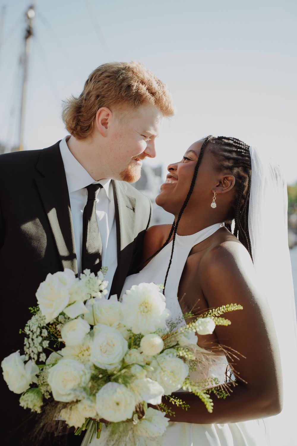 Interracial couple in wedding attire smiling at each other, bride holding white floral bouquet outdoors.