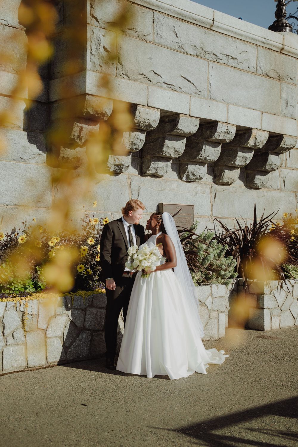 Bride and groom share a romantic moment outdoors near a stone wall with autumn flowers in the foreground.