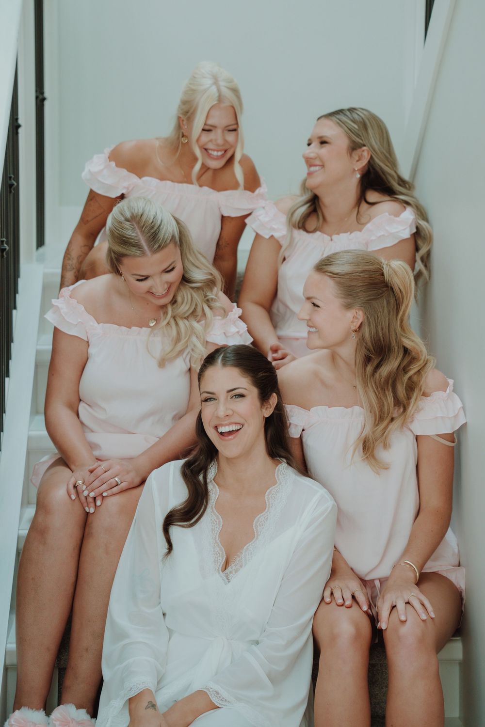 Bride in white robe laughing with four bridesmaids in pink off-shoulder dresses sitting on staircase.
