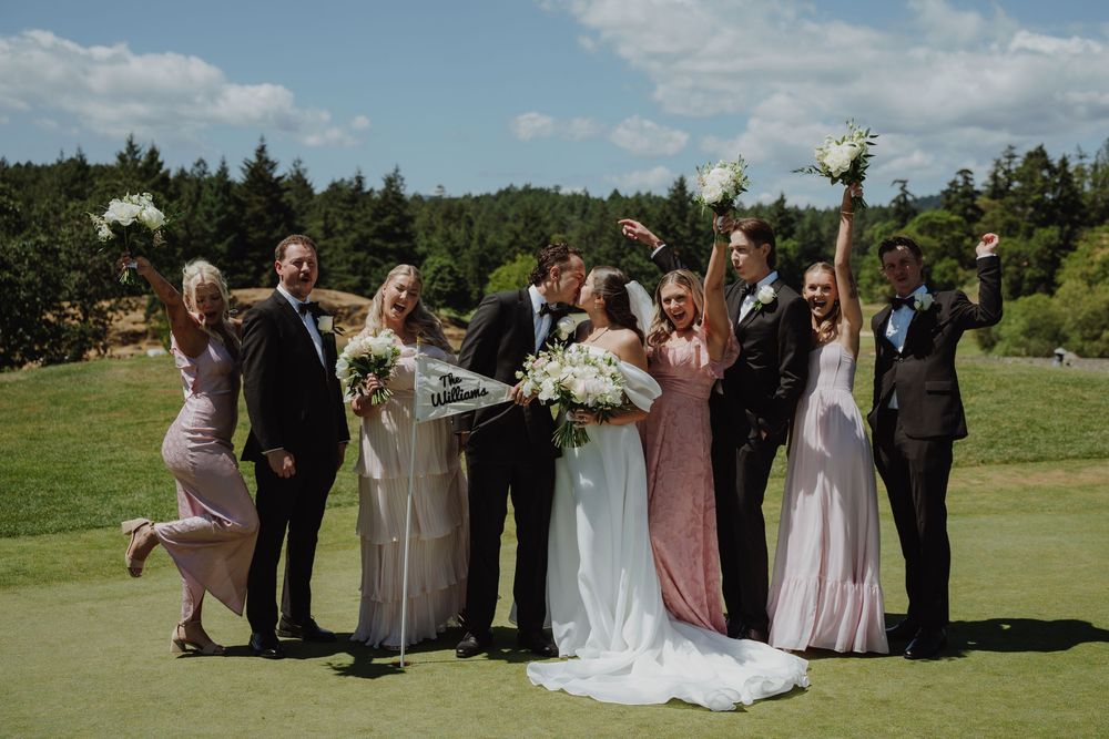 Wedding party celebrates outdoors on golf course, bride and groom kissing while bridesmaids and groomsmen cheer around them.