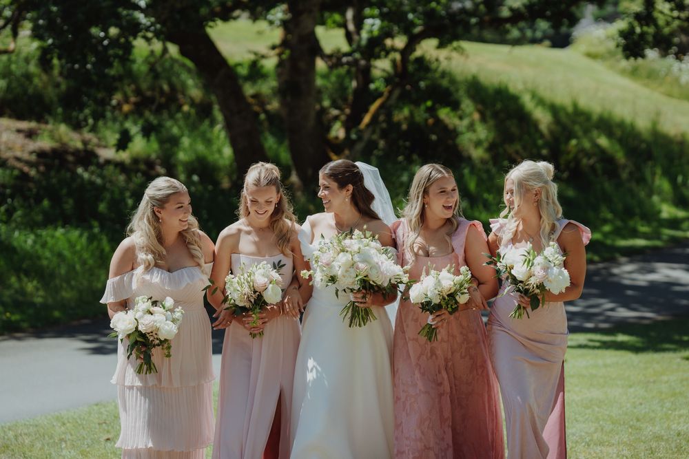Bride in white gown with bridesmaids in blush pink dresses holding white floral bouquets outdoors on a sunny day.