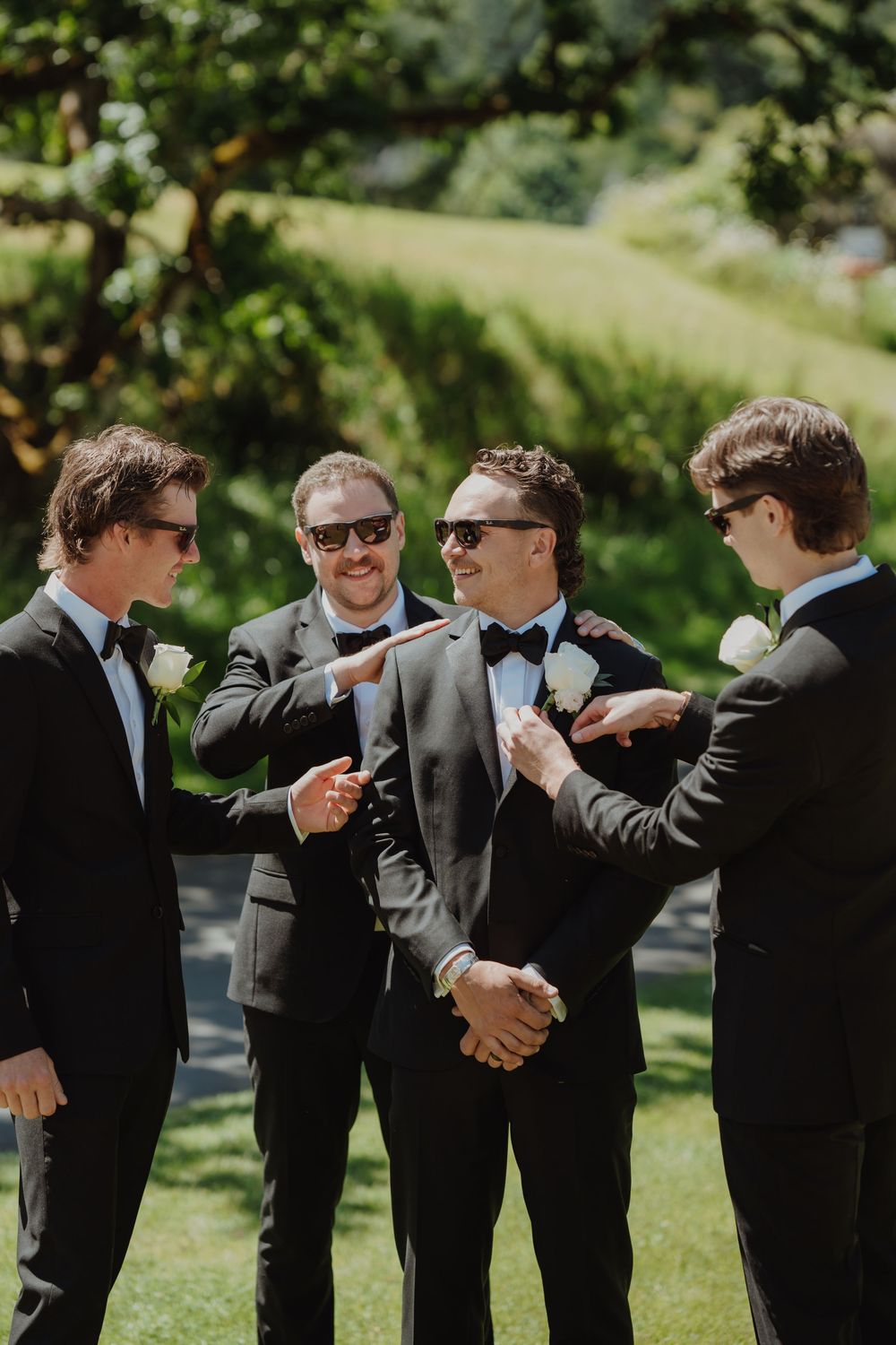 Four groomsmen in black tuxedos and sunglasses laughing outdoors, adjusting boutonnieres on a sunny wedding day.