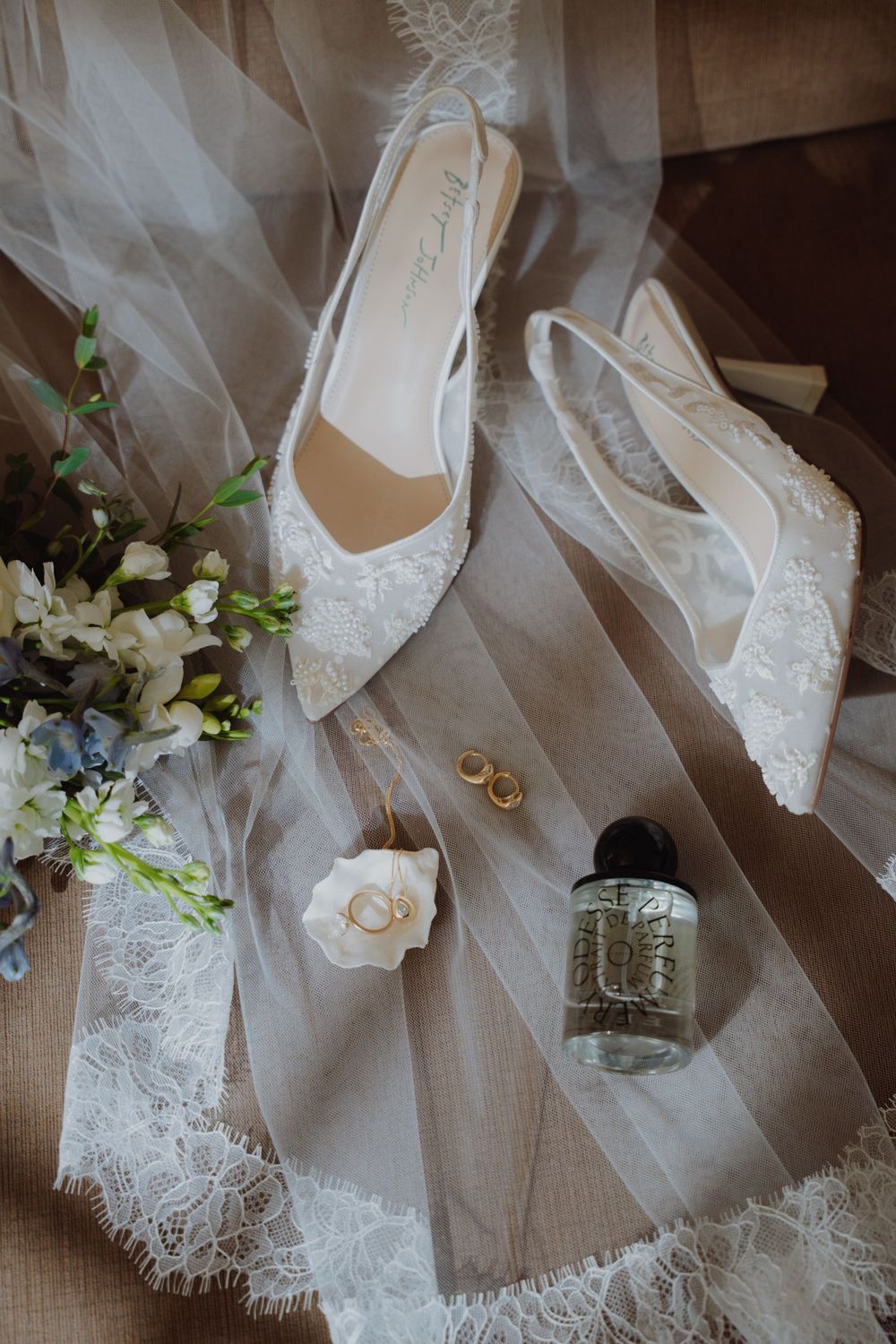 Bridal flat shoes with lace detailing displayed on tulle veil alongside gold earrings, rings, perfume, and white flowers.