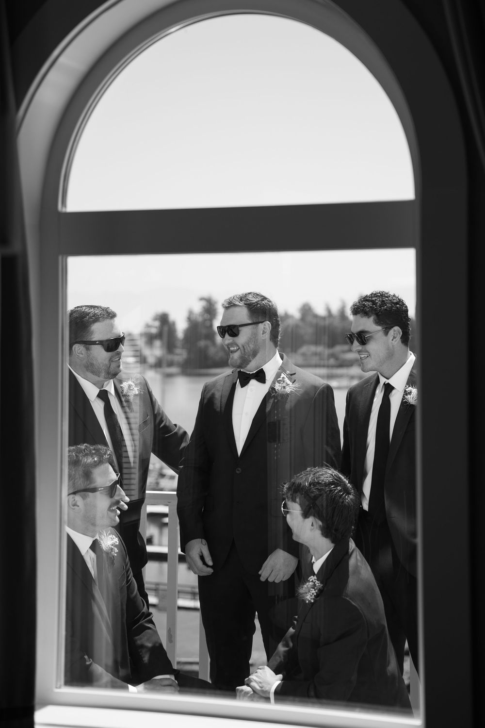 Groom and groomsmen in black tuxedos and sunglasses laughing together viewed through an arched window in black and white.