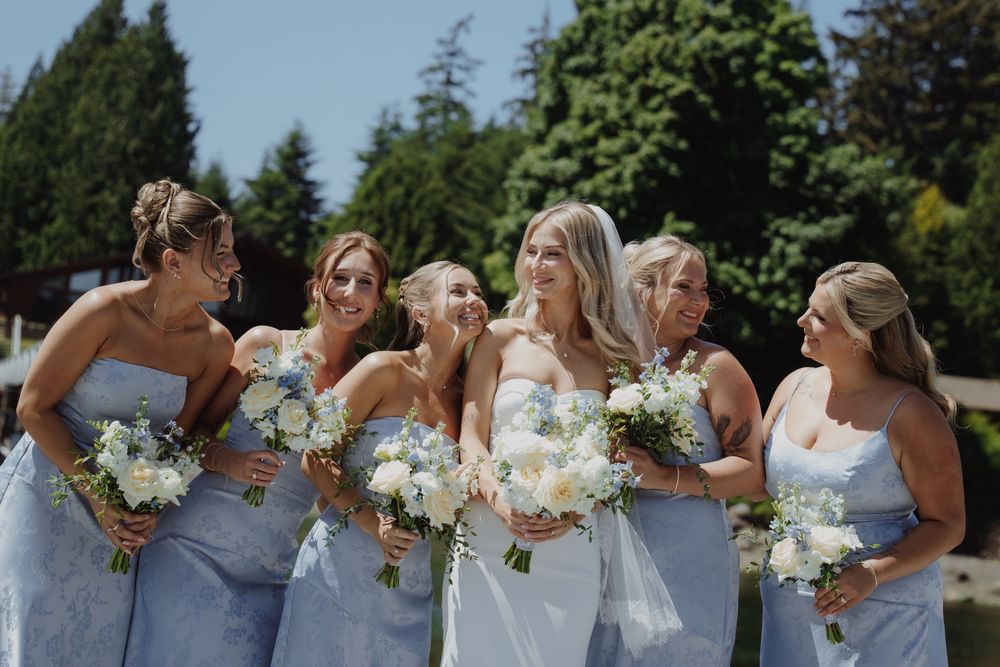 Five bridesmaids in light blue dresses laughing and holding white floral bouquets outdoors on a sunny wedding day.