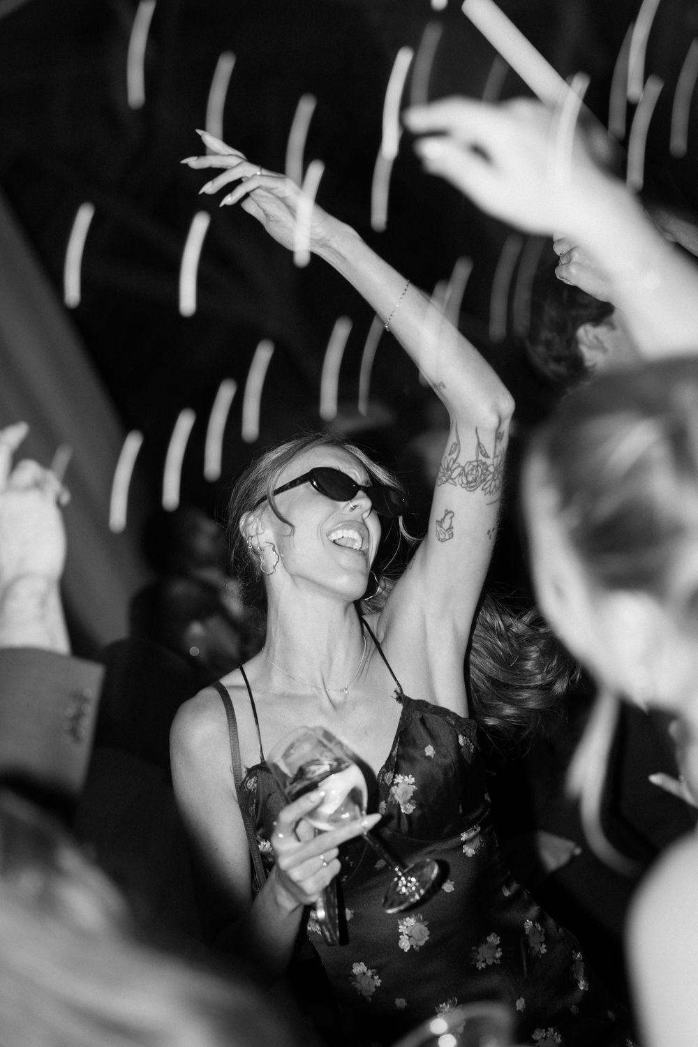 Black and white photo of a woman dancing joyfully with sunglasses and tattoos at a lively party.