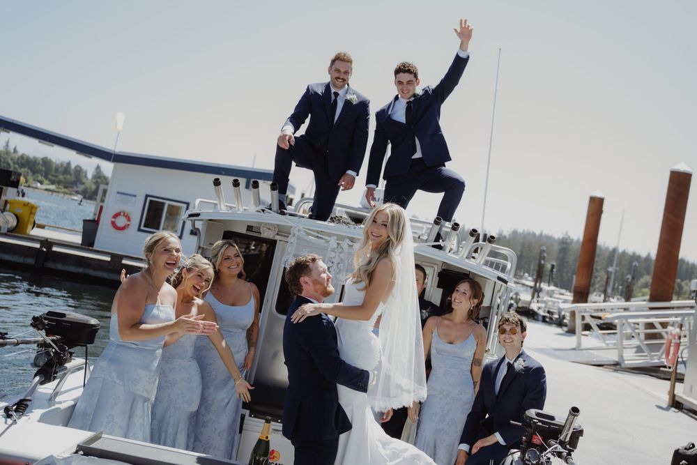 Wedding party celebrates on a boat dock, bridesmaids in blue, groomsmen cheering above, bride and groom embracing on sunny day.