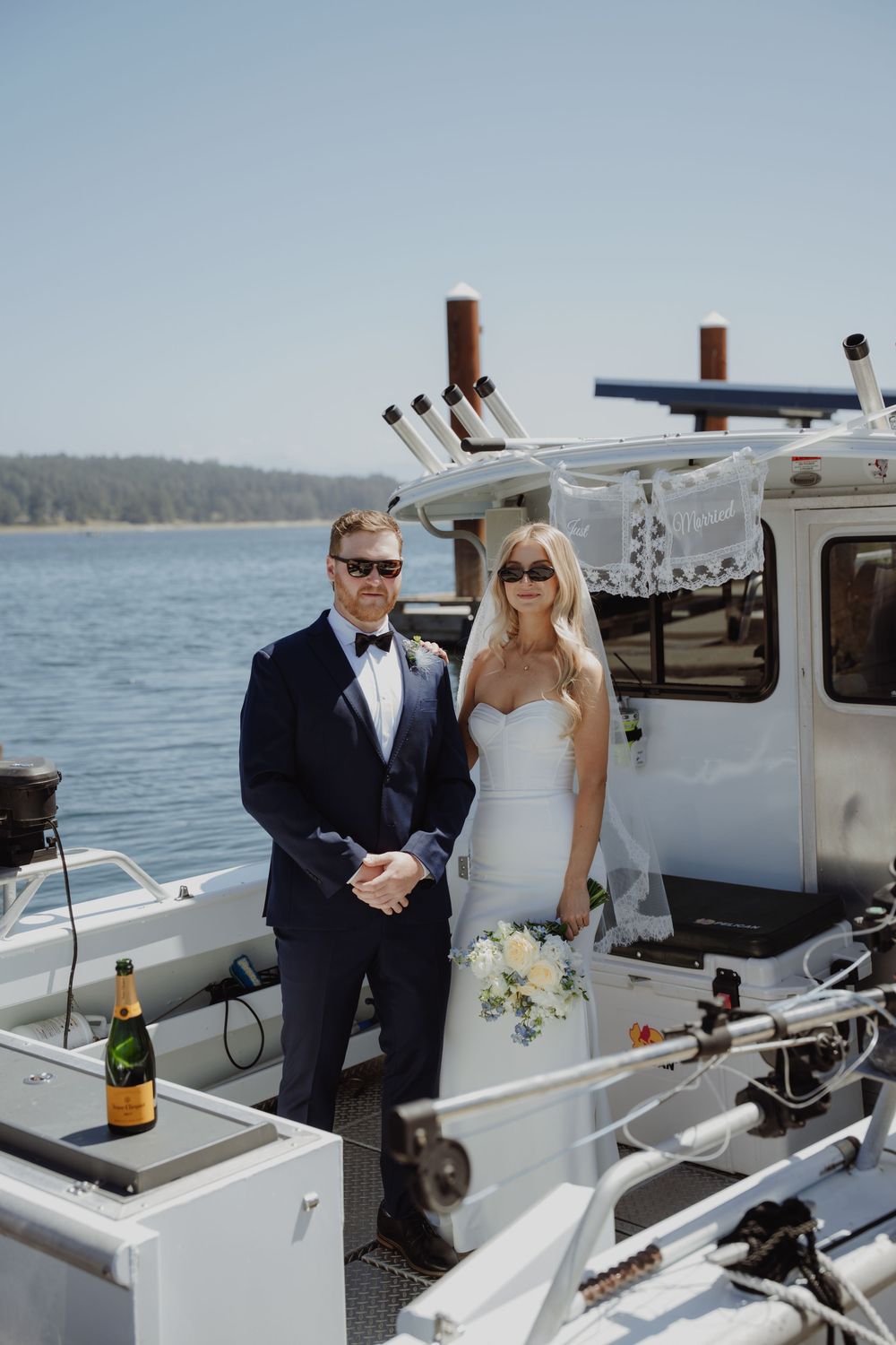 Bride and groom in wedding attire pose on a boat with champagne, water and trees in the background.