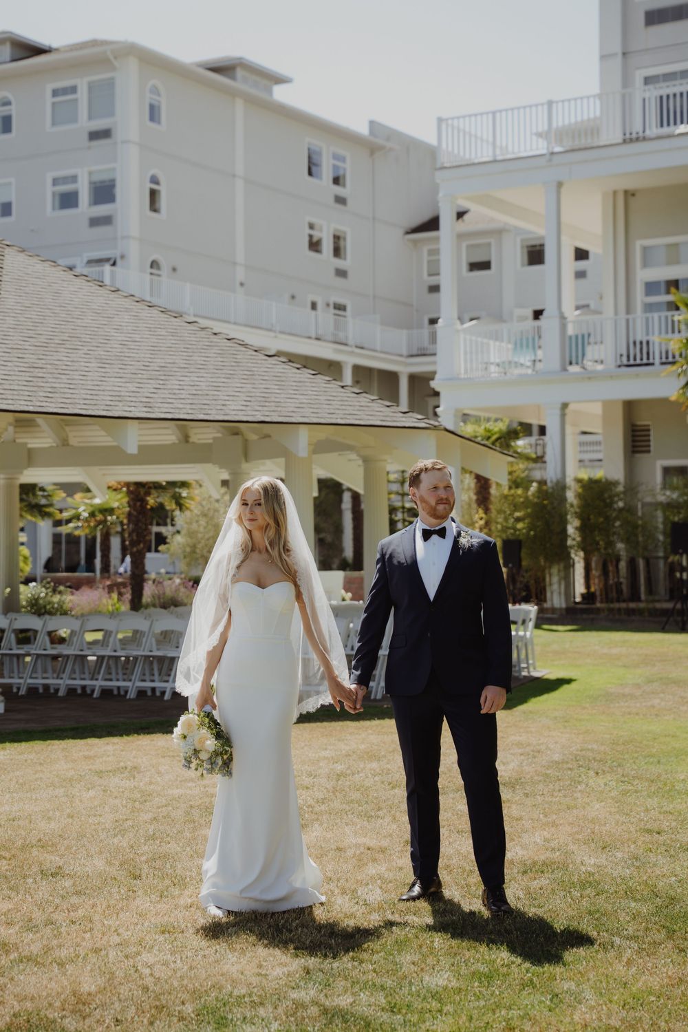 Bride in sleek white gown with veil and groom in navy tuxedo holding hands on green lawn at waterfront venue.