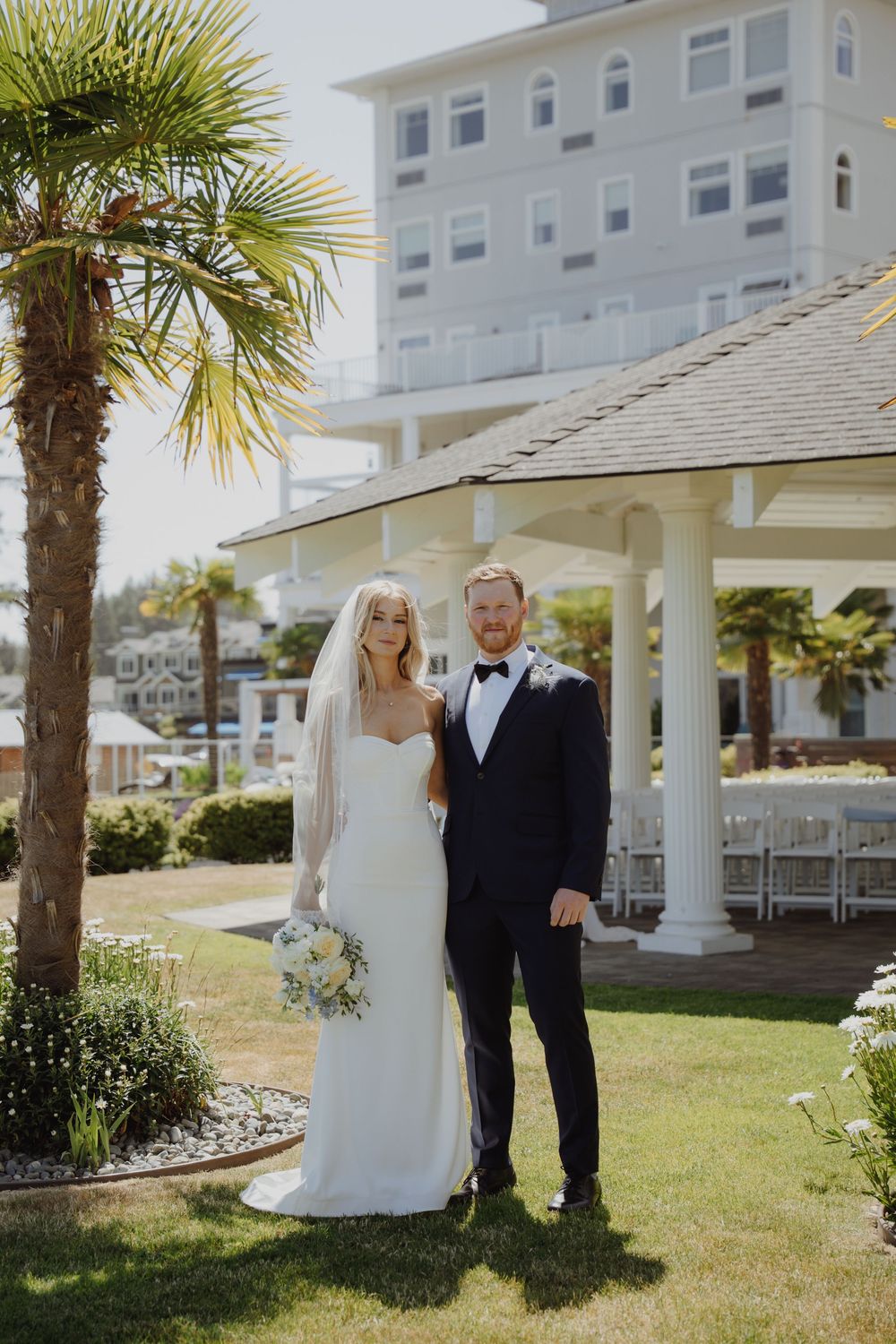 A bride and groom pose together on a sunny day outside a white coastal hotel with a gazebo and palm trees.