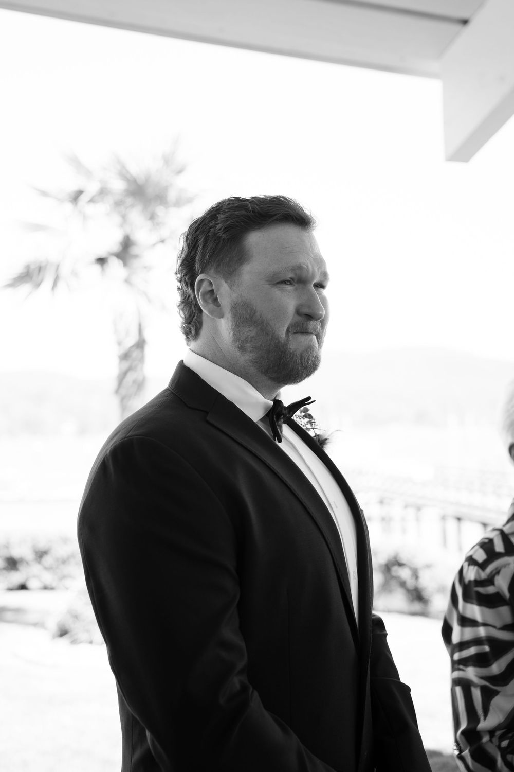 Black and white photo of a bearded groom in a tuxedo with bow tie, standing outdoors with a palm tree behind him.