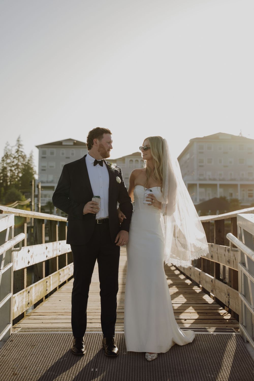 Bride and groom holding coffee cups walk on a sunny waterfront boardwalk, bride in white gown with veil.