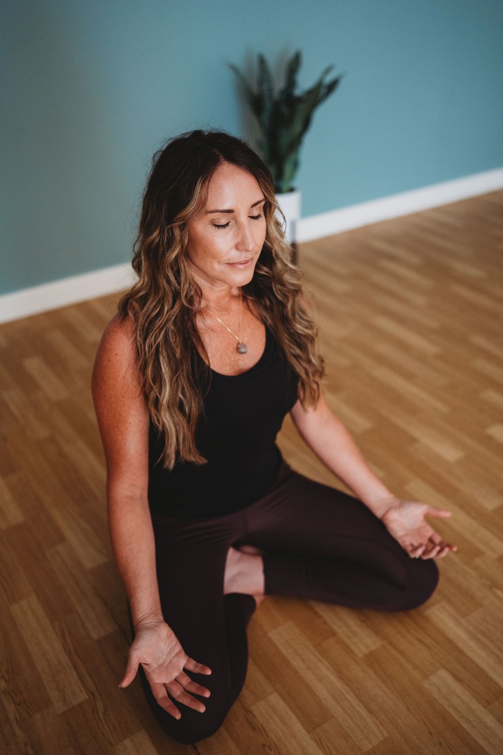 Person in black athletic wear sitting in meditation pose on hardwood floor with teal wall and plant in background.