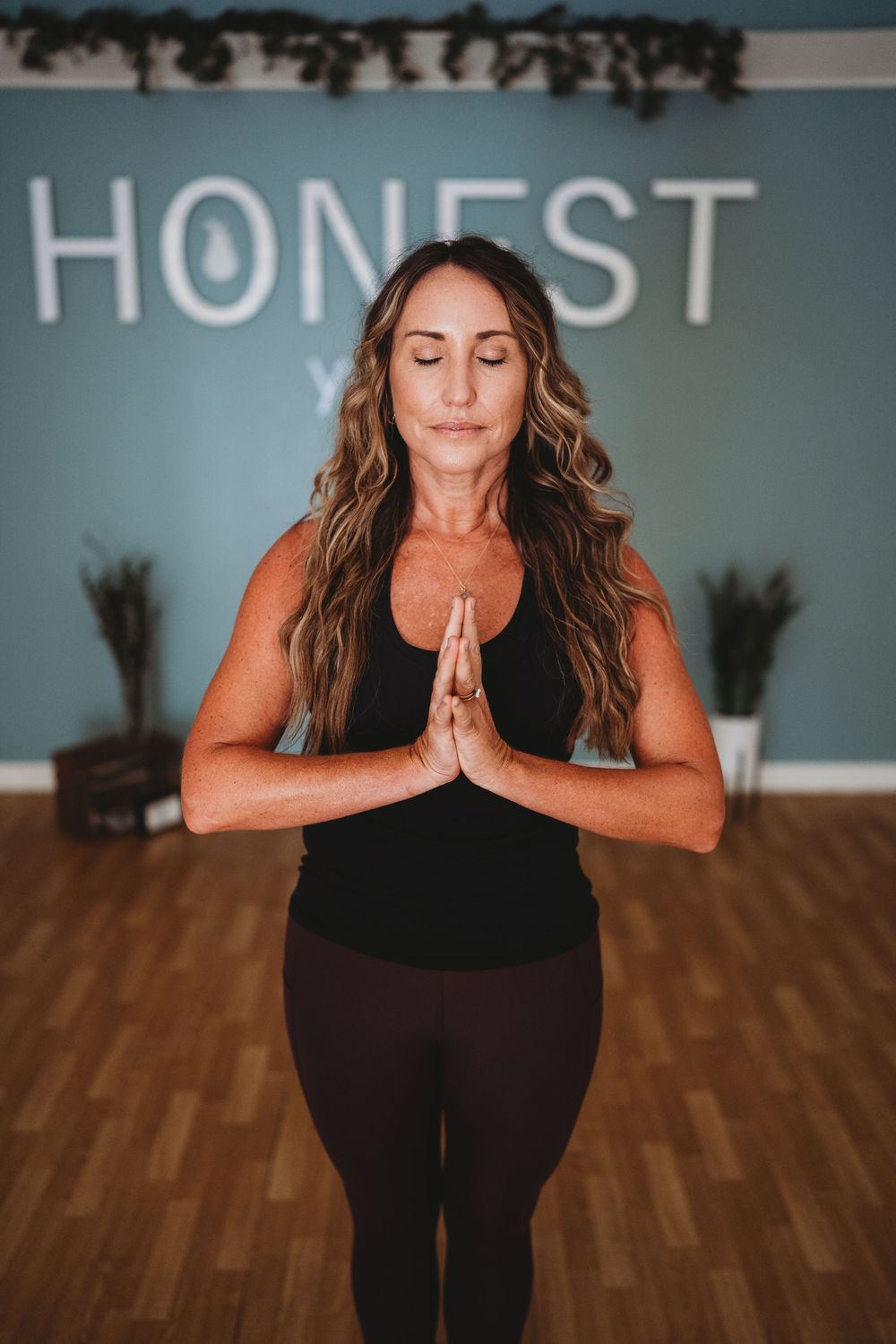 Person practicing yoga in prayer pose beneath 'HONEST' text on teal wall in yoga studio.