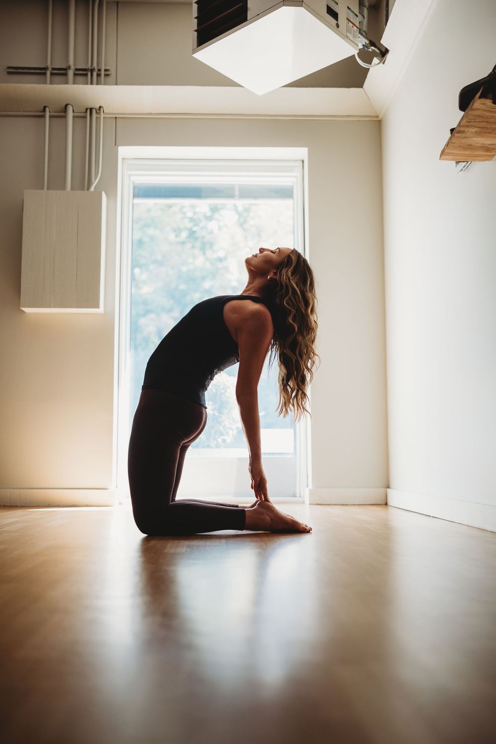 Silhouette of person performing camel pose yoga position near window in minimalist room.