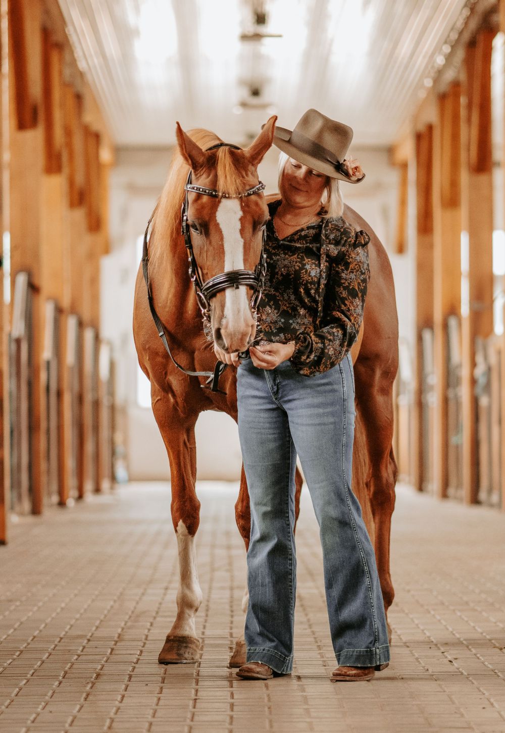 Woman in cowboy hat and floral top walking alongside a chestnut horse in a warmly lit stable aisle.
