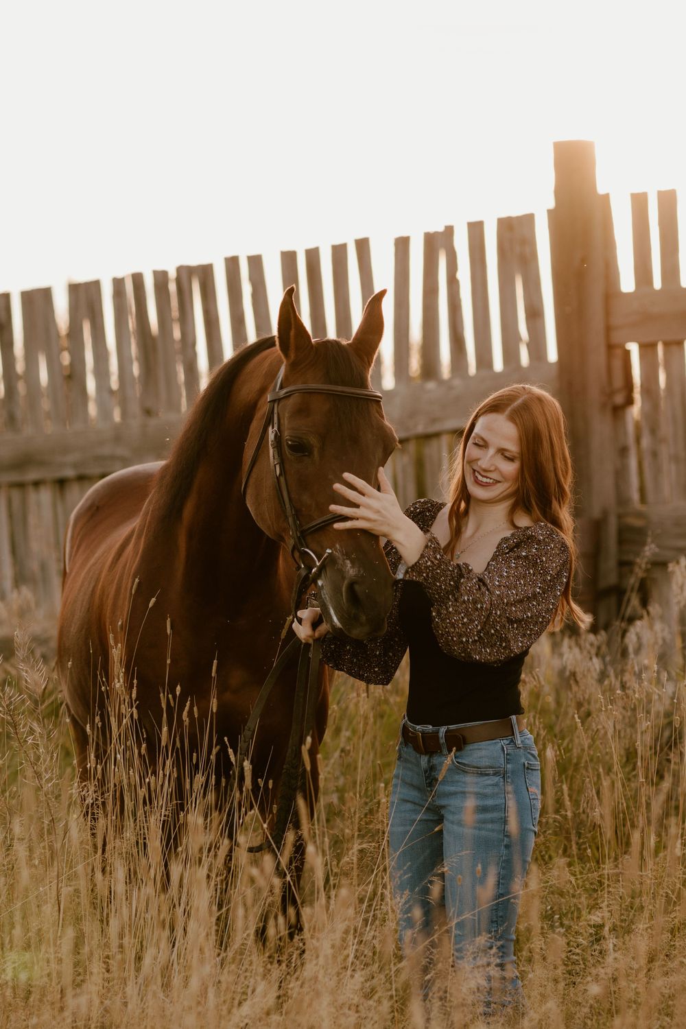 Smiling red-haired woman gently petting a dark horse at golden hour near a wooden fence.