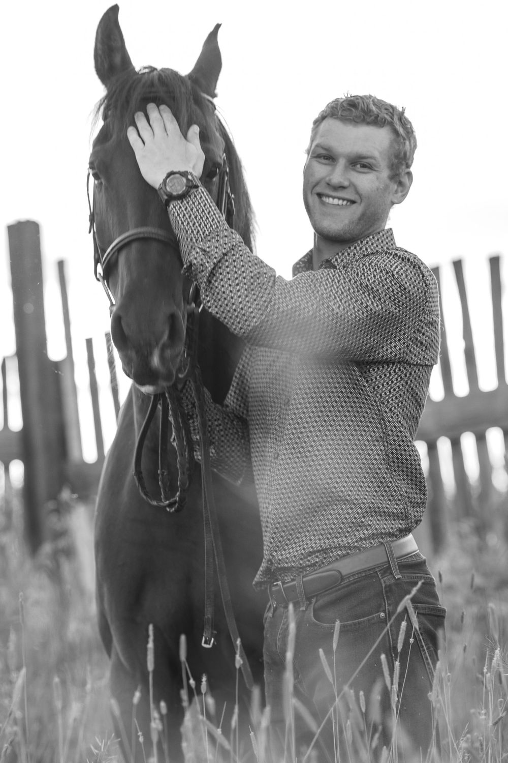 Smiling young man petting a dark horse in a field near a wooden fence in black and white.