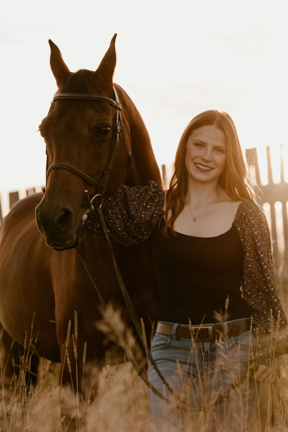 Smiling woman posing beside a chestnut horse in a golden field at sunset.