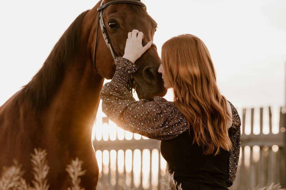 A redheaded woman affectionately holds a chestnut horse's face in glowing golden hour light.