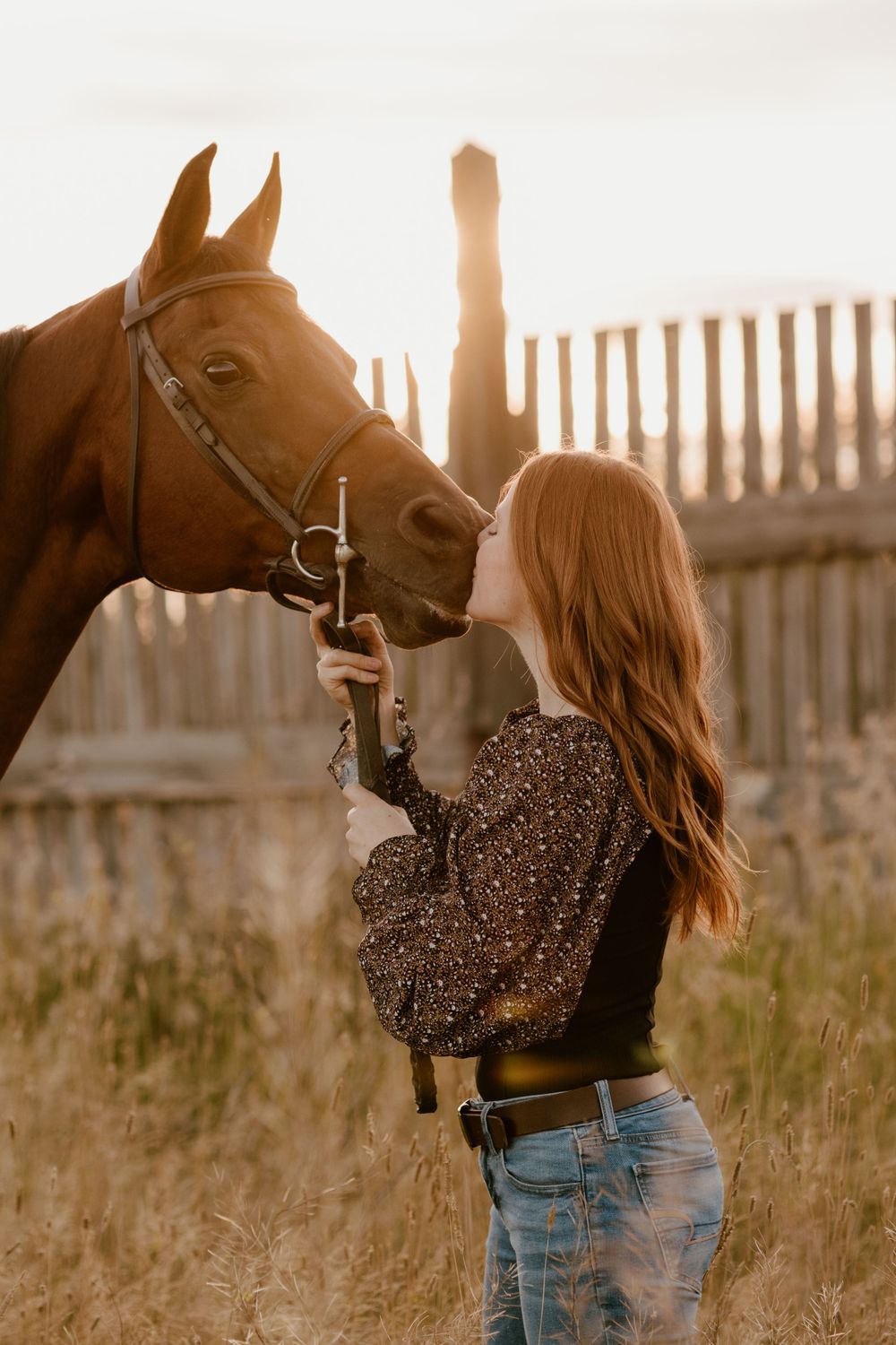 Red-haired woman kissing a chestnut horse at golden hour near a rustic wooden fence.