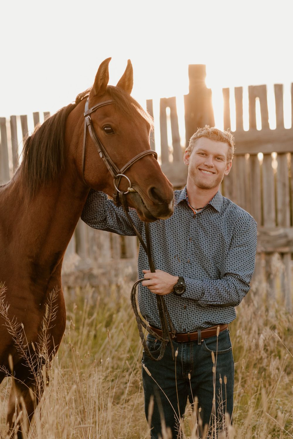 Smiling man in a blue shirt standing with a chestnut horse at sunset near a wooden fence.