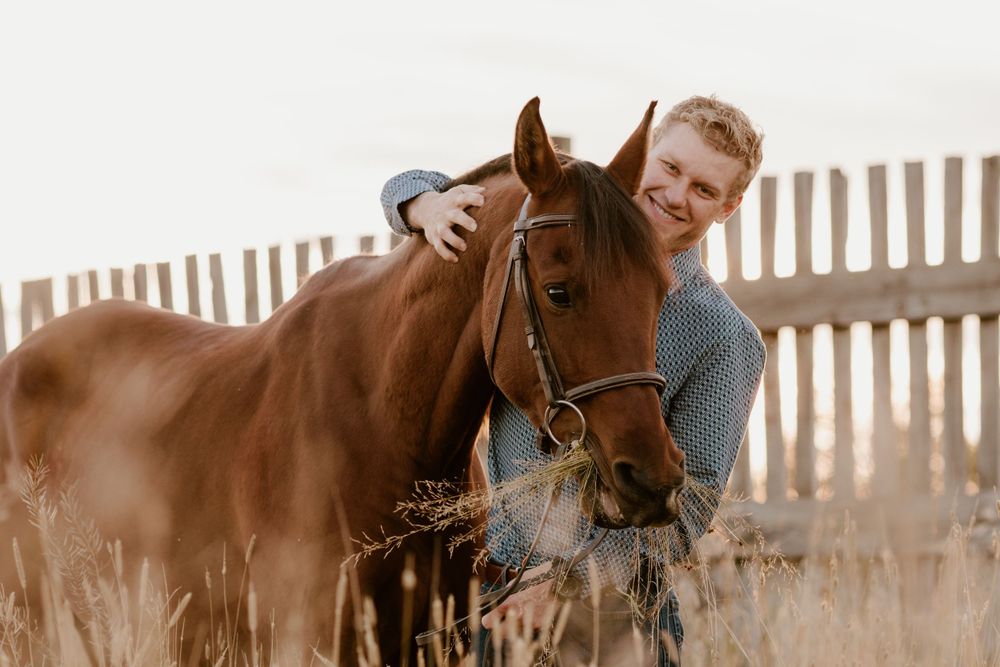 Smiling red-haired man hugging a chestnut horse in a sunny field near a wooden fence.