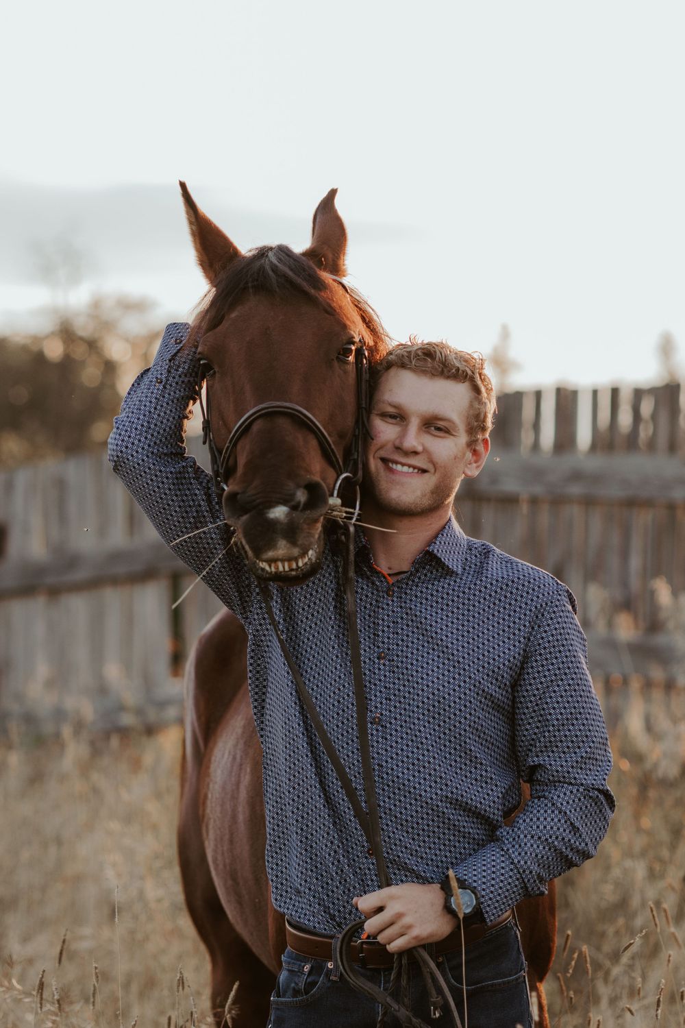 Happy man in a patterned shirt posing with a chestnut horse showing its teeth outdoors.