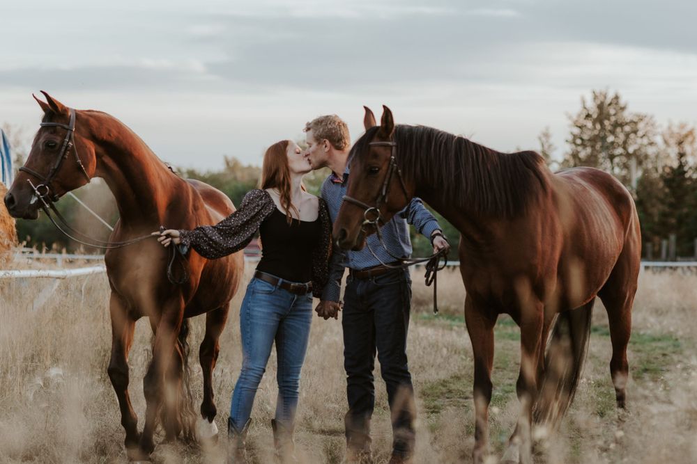 Couple kissing between two chestnut horses in an open field with autumn foliage.